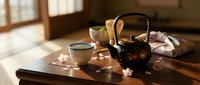 Traditional Japanese tea ceremony setup with cast iron teapot and ceramic cups on wooden table in sunlit room with cherry blossom petals for meditation and mindfulness.