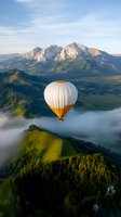 White and orange hot air balloon floating above misty mountain landscape at sunrise, with dramatic peaks and green valleys creating serene aerial scenery.
