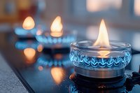 Glowing candles in decorative glass holders create cozy atmosphere with warm flames and blue ornamental pattern, soft bokeh reflections on dark surface.