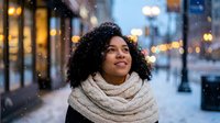 Young African American woman with curly hair wearing cozy knit scarf smiling on snowy city street with warm bokeh lights during winter evening.