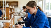 Young female scientist in blue lab coat examining sample through microscope in modern laboratory for research and scientific discovery.