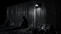 A silhouetted figure crouches beside a weathered corrugated metal structure illuminated by overhead light, evoking themes of poverty, homelessness, and urban hardship for editorial and social awareness projects.