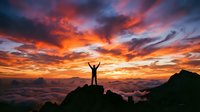 Silhouette of person with raised arms celebrating success on mountain peak at dramatic sunrise with vibrant orange and pink clouds above sea of clouds.