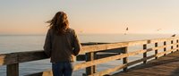 Woman with long brown hair, wearing dark jacket, standing on wooden pier at sunset gazing toward ocean horizon. Golden hour lighting, peaceful coastal scene for travel and lifestyle content.