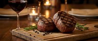 Grilled beef steaks with rosemary on wooden board paired with red wine in elegant restaurant setting with warm candlelit ambiance for fine dining experience.
