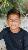 Young Hispanic boy wearing navy blue sweater smiles warmly at camera against natural green background, capturing genuine childhood joy and innocence.