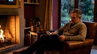 Mature Caucasian man with glasses reading book in cozy living room with fireplace. Comfortable evening at home relaxation and leisure lifestyle concept.