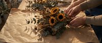 Hands arranging dried sunflowers and foliage on kraft paper in natural sunlight, perfect for floral design, crafts, and botanical styling projects.