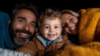 Happy diverse family with bearded father, blonde mother and curly-haired toddler boy cuddling together under cozy blankets, sharing joyful moment in dark setting.