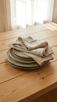 Stacked neutral ceramic plates with folded linen napkin on wooden dining table in bright sunlit room with sheer curtains, suitable for table setting and home decor.