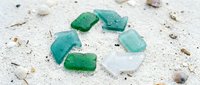 Recycling symbol made from green and white sea glass pieces arranged on sandy beach with shells and pebbles, representing environmental conservation and sustainable waste management practices.