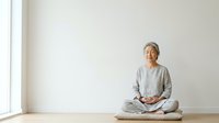 Senior woman with gray hair in casual light gray clothing sitting cross-legged on wooden floor in meditation pose against white wall with natural window light for wellness and mindfulness concepts.