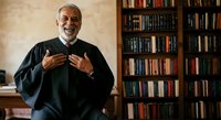 Senior male with gray beard and hair, appearing to be of Middle Eastern or Mediterranean ethnicity, wearing black judicial robes, gesturing expressively with both hands in an educational or explanatory manner, standing in front of wooden bookshelves filled with law books and legal references in a professional office or courtroom setting.