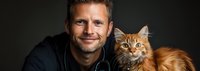 Caucasian male veterinarian in navy scrubs holding ginger Maine Coon cat against dark background, professional healthcare portrait showing trust and animal care.