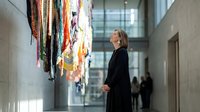 Senior woman in black coat admiring colorful textile art installation in modern museum gallery space with contemporary architecture and natural lighting.