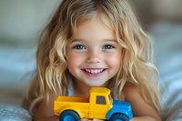 Cheerful young girl with blonde wavy hair and bright blue eyes holding yellow toy truck, showing genuine smile and joy while playing. Natural lifestyle portrait.