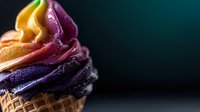 Colorful swirled soft serve ice cream in waffle cone against dark background, featuring vibrant yellow, orange, pink and purple layers with creamy texture.