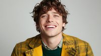 Young Caucasian man with curly brown hair wearing yellow patterned jacket and green sweater laughing genuinely against light gray background, showing white teeth and positive emotions.