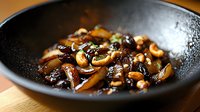 Sauteed mushrooms and cashews in dark soy sauce glaze served in black ceramic bowl, close up view of Asian fusion cuisine preparation.
