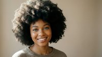 Young African American woman with natural curly afro hair smiling warmly at camera wearing beige sweater against neutral background for lifestyle content.