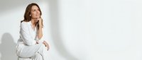 Professional middle-aged Caucasian businesswoman in white suit sitting thoughtfully against minimalist background for corporate presentations.