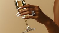 Elegant dark-skinned female hand with silver rings and white manicure holding champagne glass against beige background, luxury lifestyle concept.