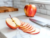 Fresh red apple sliced on white marble countertop with kitchen knife, showing cross-section and thin circular cuts, bright natural lighting in modern kitchen setting.