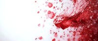 Red liquid splash with dynamic droplets and bubbles creating abstract motion effect against white background, high-speed photography capture for beverage advertising.