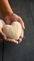 Hands holding heart-shaped dough with flour on dark wooden surface for baking and cooking projects.