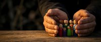 Close-up of hands protecting colorful wooden peg dolls representing diverse people, symbolizing care, community support, family protection and social responsibility concepts.