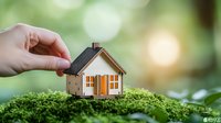 Small wooden house model placed on green moss with hand holding roof, soft bokeh background, representing eco-friendly living, real estate, or sustainable housing concept.
