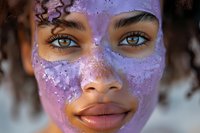 Young African American woman with natural curly hair wearing purple glitter face mask, close up portrait showing bright blue eyes and gentle smile. Spa and skincare concept.