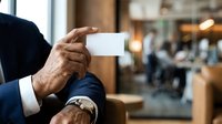 Mature male professional in dark business suit holding blank white business card with wrinkled hands and wristwatch, indoor office or corporate environment with blurred background.