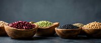 Wooden bowls filled with assorted legumes and pulses including red kidney beans, green lentils, black lentils, and chickpeas arranged on dark surface for cooking and nutrition content.