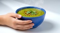 Woman hands holding blue ceramic bowl with green pea soup garnished with herbs and spices on white background for healthy eating and cooking concepts.