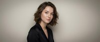 Professional young Caucasian businesswoman with curly brown hair in black blazer against neutral background for corporate marketing materials.