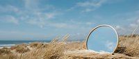 Round mirror with golden frame standing among dry beach grass dunes under blue sky, reflecting ocean horizon, summer coastal scene.