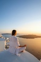 Woman in white robe meditating at sunset overlooking ocean and mountains from Mediterranean terrace during golden hour wellness retreat.