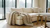 Cozy modern living room with cream sectional sofa, knitted throw blanket, wooden side table, and large windows with natural light for home decor and interior design projects.
