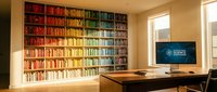 Modern home office with colorful rainbow bookshelf wall featuring organized books from red to blue spectrum, wooden desk and computer setup for remote work productivity.