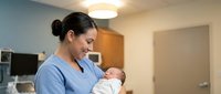 Female healthcare worker in blue scrubs holding newborn infant in hospital room setting, maternity care and nursing.