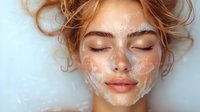 Young Caucasian woman with closed eyes and messy blonde hair during skincare routine, face covered in white foam cleanser against light background.