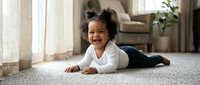 Happy African American toddler girl crawling on carpet in bright living room with natural sunlight streaming through windows creating warm family atmosphere.