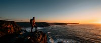 Young man standing on rocky coastal cliff at golden hour sunset, watching ocean waves with scenic landscape backdrop for travel and lifestyle content.