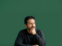 Middle-aged man with dark hair and facial hair, appearing thoughtful and contemplative with hand near face against green background for portrait use.