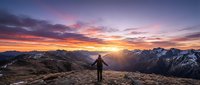 Hiker standing on mountain summit at sunrise with golden sun rays breaking through purple and orange clouds over alpine peaks.
