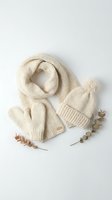 Cozy cream knitted winter accessories set with scarf, beanie hat and mittens arranged on white background with eucalyptus branches for seasonal fashion styling.