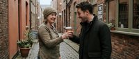 Two young adults, one wearing a beige knit beanie and olive jacket, the other in a black coat, exchange a warm beverage while standing in a charming brick alley. Urban street encounter.