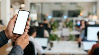 Hand holding smartphone with blank white screen in modern office environment with blurred coworkers and workspace in background.