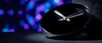 Modern black round clock with silver hands on dark surface against colorful bokeh lights background for time management and business concepts.